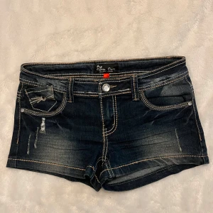 Mörkblå jeansshorts med broderade fickor - Snygga lågmidjade jeansshorts med slitningar och dekorativa sömmar på bakfickorna. De har en knapp och dragkedja framtill. Skriv ifall du har några frågor. 