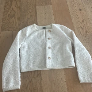 Vit bouclé kofta med guldknappar - Denna kofta ger en väldigt elegant look på outfiten och är superfin över ett vanligt linne. Guldknapprna är en väldigt fin detalj. Koftan är mjuk och mysig, har en texturerad yta. Den kommer från Gina Tricot och är i storlek S.