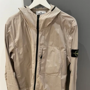 Äkta Stone Island jacka - Snygg beige vår/höstjacka från Stone Island med huva och dragkedja. Jackan har en ficka på bröstet och den ikoniska logotypen på ärmen. Passar perfekt till våren eller hösten. Original pris 6000kr säljer för 3500. Nyskick bara använd ett fåtal ggr. 