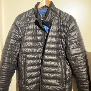  Prada Jacka  - Snygg svart quiltad jacka från Prada med dragkedja och två fickor framtill. Jackan har en stilren design och är perfekt för kyligare dagar. Insidan är fodrad med blått tyg som ger en fin kontrast.