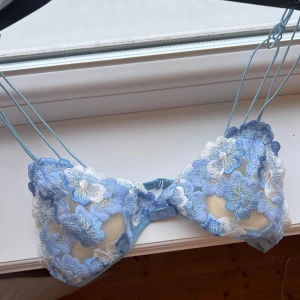Blå blommig spets-bh - Säljer en vacker blå bh med blommigt spetsmönster. Den har tunna axelband och en delikat design med vita och blå blommor. Perfekt för att känna sig extra fin. Aldrig använd 