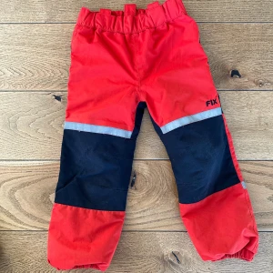 Röda byxor från Fix - Säljer ett par röda byxor från Fix med svarta detaljer på knäna och reflexband. Perfekta för utomhusaktiviteter med elastisk midja för bekväm passform.
