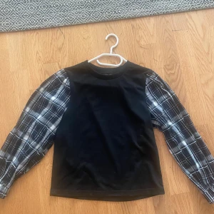 Svart tröja med rutiga ärmar  - Snygg svart tröja från Shein med rutiga ärmar i grått och vitt. Tröjan har en normal passform och rund halsringning.