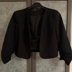 Svart kavaj från FB sister - Elegant svart kavaj från FB sister med en stilren design. Kavajen har halvlånga ärmar med rynkade detaljer vid ärmsluten, vilket ger en unik touch. Perfekt för att komplettera en chic outfit. Innertyget i ärmslutet på båda ärmarna har gått sönder. Inget som syns och inget som påverkar användandet. Se bilder.