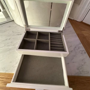Vitt smyckeskrin - Elegant vitt smyckeskrin med glas i locket. Skrinet har flera fack för smycken och en utdragbar låda för extra förvaring. Perfekt för att organisera dina smycken på ett stilfullt sätt.