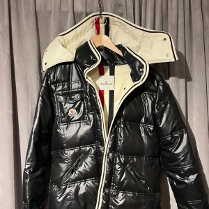 Svart dunjacka stl S - Snygg svart dunjacka från Moncler med vit insida och detaljer. Jackan har en hög krage och praktiska fickor med knappar. Perfekt för kyliga dagar med sin stilrena design och varma material.  En av tryckknapparna har lossnat men det stör inte funktionen.