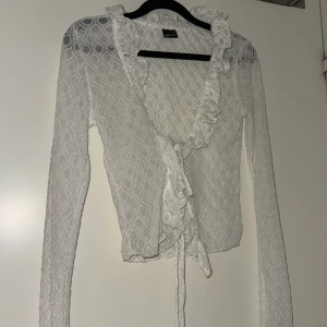 Vit spetsblus med volangdetaljer - Elegant vit spetsblus från Ginatricot med vackra volangdetaljer och knytning framtill. Blusen har ett diskret mönster och långa ärmar, perfekt för en stilren look.