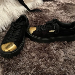 Svarta sneakers från Puma med gulddetaljer - Snygga svarta sneakers från Puma med en iögonfallande gulddetalj på tån och guldfärgad logga. Skorna har snörning och en robust sula som ger extra komfort.