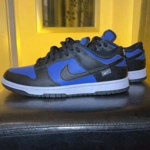 Nike Dunk i svart och blått - Snygga Nike Dunk sneakers i svart och blått med vit sula. Skorna har klassisk snörning och en stilren design med Nike-logga på hälen. Perfekta för en sportig look.