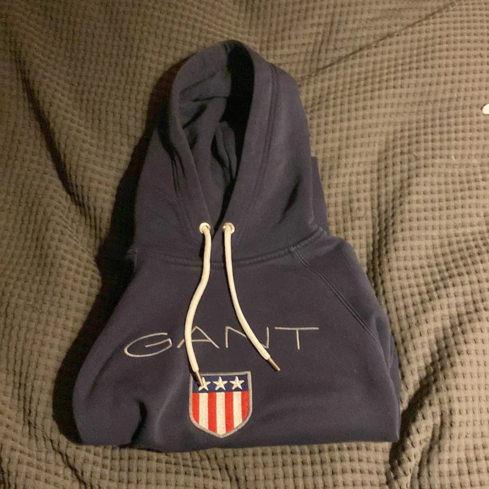 Säljer en snygg mörkblå hoodie från GANT med vit dragsko och ett coolt emblem på bröstet. Använd få gånger. Perfekt för en avslappnad stil. Hör av dig vid intresse!. Neuletakit & Villapaidat.