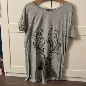 Grå t-shirt med hjortmotiv från Lindex - Säljer en grå t-shirt från Lindex med ett stort svart hjortmotiv på framsidan. T-shirten har korta ärmar och en rund halsringning. Kan användas som nattlinne har aldrig använts