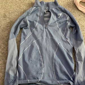 Blå långärmad tröja från Gymshark - Säljer en blå långärmad tröja från Gymshark med dragkedja vid halsen och vita detaljer. Perfekt för träning eller en aktiv vardag. Tröjan har en praktisk ficka med dragkedja på ryggen.