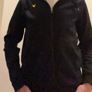 Svart huvtröja med dragkedja från Lyle & Scott - Snygg svart huvtröja från Lyle & Scott med dragkedja och gul logga på bröstet. Perfekt för en casual look. Tröjan har långa ärmar och en bekväm huva.