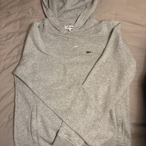 Grå hoodie från Lacoste Sport - Säljer en stilren grå hoodie från Lacoste Sport med den klassiska krokodilloggan på bröstet. Tröjan har en bekväm passform och är perfekt för en avslappnad look. Den har en justerbar huva och ribbade muddar. Stl 176 ca 15 år.  Något liten i stl. Endast använd en gång. 