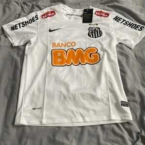 Neymar Santos från Nike - Säljer en santos tröja med Neymar från Nike med korta ärmar och tryck från Banco BMG och Netshoes. Tröjan har Dri-FIT teknologi för att hålla dig torr och bekväm. Perfekt för fotbollsfans!