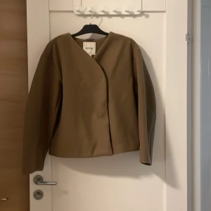 Brun jacka från Monki - Säljer en stilren beige jacka från Monki i storlek M. Jackan har en minimalistisk design med tryckknappar framtill och långa ärmar. Perfekt för en chic och enkel look.