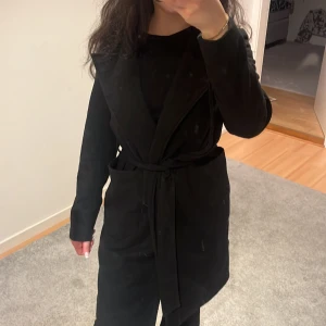 Svart kappa med knytband - Elegant svart kappa med knytband i midjan. Perfekt för en stilren look. Långärmad design med klassisk passform. Den är ifrån Noisy may, storlek S, pris kan diskuteras 