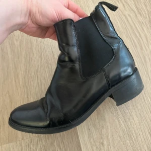 Svarta boots från Tiger of Sweden - Svarta Chelsea boots i skinn från Tiger of Sweden. Klassisk design med en liten klack. Finns spår av användning (finns ett litet ”hack” i ena skon se bild) men annars i gott skick. 
