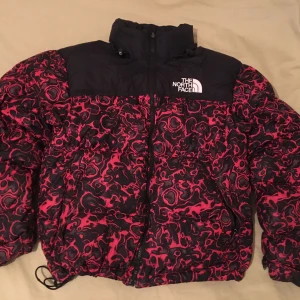 North Face 1996 Retro Nuptse Jacket Rose Red '94 Rage Print - Snygg pufferjacka från The North Face i svart och rött med unikt mönster. Jackan har en hög krage och dragkedja framtill. Perfekt för kyliga dagar.