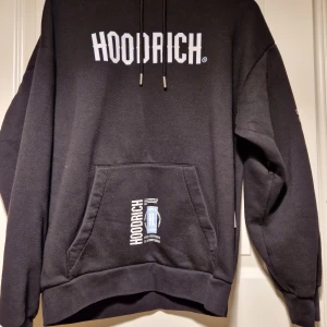 Svart hoodie från Hoodrich - Snygg svart hoodie från Hoodrich med vit logga på bröstet och detaljerad text på ryggen. Den har en stor ficka framtill och justerbar huva med dragsko. Perfekt för en avslappnad stil.