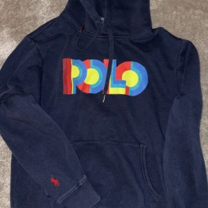 Mörkblå hoodie från Polo Ralph Lauren - Säljer en snygg mörkblå hoodie från Polo Ralph Lauren med färgglatt tryck på bröstet. Tröjan har en klassisk känguruficka och justerbar huva. Perfekt för en avslappnad stil.