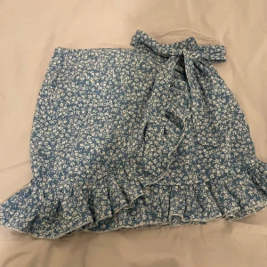 Blommig kjol från Shein - Charmig blå blommig kjol från Shein med volangdetaljer och knytning i midjan. Perfekt för en somrig look med sitt lätta och luftiga material.