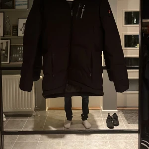 Svart dunjacka från Sail Racing - Säljer en svart dunjacka från Sail Racing med broderad logga och texten 'Sail Antarctica Expedition'. Jackan har en långärmad design och dragkedja framtill. Perfekt för kalla dagar med sin varma och skyddande stil.