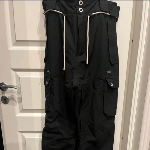 Svarta byxor från Dope - Säljer ett par svarta byxor från Dope med praktiska fickor och snörning i midjan. Perfekta för utomhusaktiviteter med stilren design.