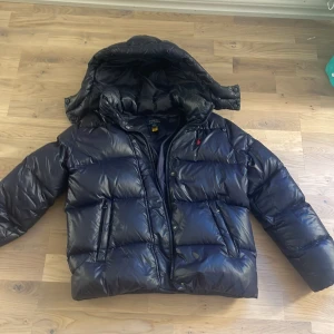 Marinblå dunjacka från Polo Ralph Lauren - Säljer en svart dunjacka från Polo Ralph Lauren med luva och dragkedja. Jackan har en pufferdesign som ger extra värme och stil. Perfekt för kyliga dagar.