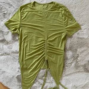 Grön kortärmad topp med snörning M/L - Snygg grön topp med korta ärmar och trendig snörning framtill som ger en rynkad effekt. Perfekt för en avslappnad stil med en twist. Tillverkad i ett mjukt och bekvämt material.