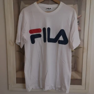 Vit t-shirt från FILA - Säljer en klassisk vit t-shirt från FILA med stor logga i blått och rött på framsidan. T-shirten har korta ärmar och rund halsringning.
