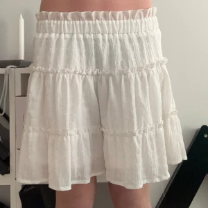 Vit volangkjol - Charmig vit kortkjol med volanger. Perfekt för sommarens varma dagar. Den elastiska midjan ger en bekväm passform och det luftiga materialet gör den sval och skön att bära. Storlek S men den elastiska midjan kan annpassa storleken.