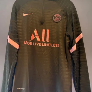 Psg långärmad fotbollströja från Nike - Snygg svart långärmad fotbollströja från Nike med rosa detaljer och PSG-logga. Perfekt för fotbollsfans som vill ha en stilren look. Tröjan har en dragkedja vid halsen och Dri-FIT-teknologi för optimal komfort.