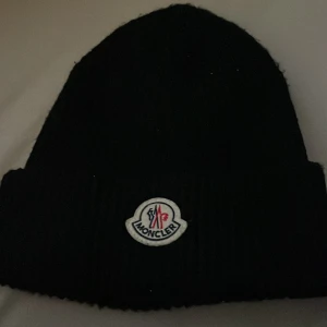 Svart mössa från Moncler - Snygg svart mössa från Moncler med ribbad design och det klassiska Moncler-emblemet framtill. Perfekt för att hålla värmen under kyliga dagar.