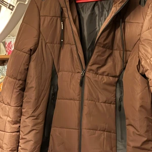 Brun pufferjacka från Bluewear - Säljer en brun pufferjacka från Bluewear med dragkedja och justerbar huva. Jackan har svarta detaljer på axlarna och är perfekt för kalla dagar. Den är långärmad och har en quiltad design för extra värme.