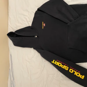 Mörkblå hoodie från Polo Sport Ralph Lauren - Säljer en mörkblå hoodie från Polo Sport Ralph Lauren med broderad logga i gult och rött samt en amerikansk flagga på bröstet. Hoodien har en klassisk design med långa ärmar och en stor ficka framtill. Hoodien har även en snygg logga på högerarmen. Hoodien är en L i barn storlek. Den passar vanligt S/M. Det finns en QR-kod i nacken som leder till ralphlauren.eu där det visar att den e äkta. Finns det några frågor angående passform skriv bara till mig      #grisch#laidback#ralphlauren#polo#hoodie