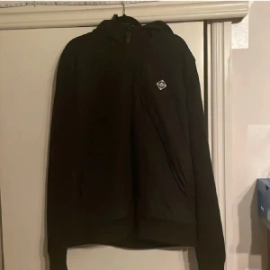 Svart jacka från J.Lindeberg - Snygg svart jacka från J.Lindeberg med en stilren design. Jackan har en huva och en diskret logotyp på bröstet. Perfekt för en modern och minimalistisk look. Sitter som L/M