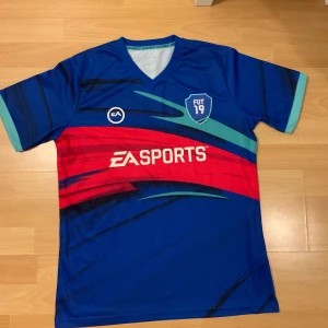 FIFA 19 tröja - Tröja från FIFA 19, bra skick och fina färger. Det enda problemet med det är EA sport trycket som har börjat falla av, syns på bilden men är inte så märkvärdigt. 