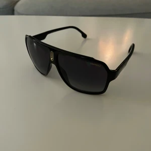 Svarta solglasögon från Carrera - Snygga svarta solglasögon från Carrera med en sportig design. De har en bred ram och mörka linser som ger ett coolt och stilrent intryck. Perfekta för soliga dagar! Nypris 1400kr