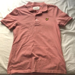 Rosa pikétröja från Lyle & Scott - Snygg rosa pikétröja från Lyle & Scott med klassisk krage och knappslå. Tröjan har en broderad logotyp på bröstet och är tillverkad i ekologiskt material. Perfekt för en fest stil!