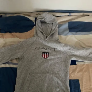 Grå hoodie från GANT - Säljer en stilren grå hoodie från GANT med logga och emblem på bröstet. Tröjan har en klassisk känguruficka och en bekväm huva. Pris kan diskuteras.