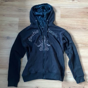 Svart hoodie från Blinglucid - Snygg svart rhinestone hoodie från Blinglucid med dragkedja. Texten 'Guardian Angels' pryder framsidan tillsammans med ett coolt mönster. Alla rhinestones sitter kvar och tröjan har blivit tvättad för hand 1 gång. Bra skick på utsidan och helt okej skick på insidan. Storlek S. Nypris Ca 900kr, kan säljas för billigare vid snabb affär.