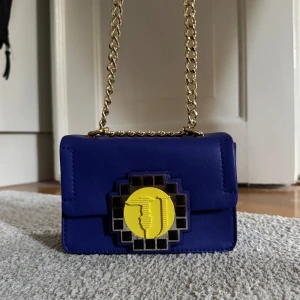 Blå trussardi axelväska med kedja - Snygg blå axelväska med gulddetaljer och en unik gul och svart dekor på framsidan. Väskan har en kedjerem och är perfekt för att ge en färgklick till din outfit. Den är som helt ny och är aldrig använd