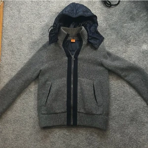 Cardigan - Snygg stickad grå cardigan med avtagbar luva från hugo boss. Mörkblått dun på rygg och luva, den är i mycket bra skick. Det är bara att skriva om ni undrar något.(Pris är diskuterbart vid snabb affär)