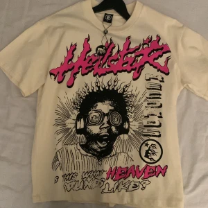 Vit t-shirt med tryck från Hellstar - En äkta och snygg vit t-shirt från Hellstar med ett stort grafiskt tryck i svart och rosa på framsidan. Trycket föreställer en person med hörlurar och texten 'hellstar'. Baksidan har också ett tryck med texten 'Hell star Records'. Perfekt för dig som gillar unika och uttrycksfulla plagg. Tagg finns kvar