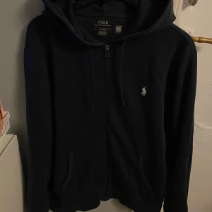 Marinblå hoodie från Polo Ralph Lauren - Snygg marinblå hoodie från Polo Ralph Lauren med dragkedja och klassisk logga på bröstet. Perfekt för en avslappnad stil. Den har en justerbar huva och praktiska fickor framtill. INTRESSE KOLL