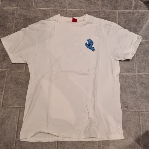 Vit t-shirt med blått tryck - Säljer en vit t-shirt med ett coolt blått tryck av en skrikande hand på både fram- och baksidan. Perfekt för en avslappnad stil. Märke Santa Cruz. Knappt använd. Storlek L