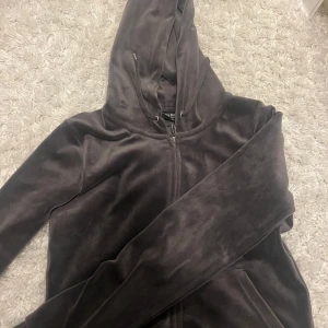 Mörkgråa hoodie från Juicy Couture - Säljer min hoodie från Juicy Couture med dragkedja och huva. Den här snygga dragkedjan genom huvtröjan är gjord av mjuk velour. Den har en klassisk passform och en bekväm känsla. Huvtröjan har en huva med dragsko, en fullängdsdragkedja och ribbade muddar och nederkant. Den är perfekt för avslappnad användning och kan stylas med jeans eller leggings. Den är i storlek XS och har en klassisk Juicy Couture-design.