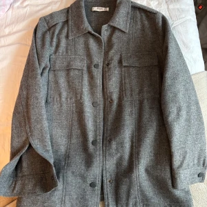 Grå ulljacka från Mango - Snygg grå ulljacka från Mango med knappar framtill och två bröstfickor. Jackan har en klassisk krage och långa ärmar.