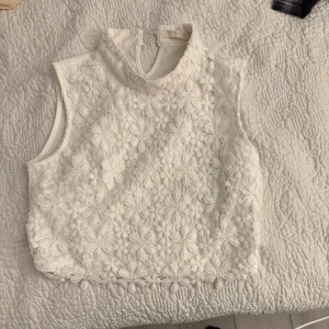 Vit spetsblus - Säljer en elegant vit spetsblus från Vera & Lucy. Blusen är ärmlös med ett vackert blommönster och knäppning i nacken. Perfekt för en stilren look.